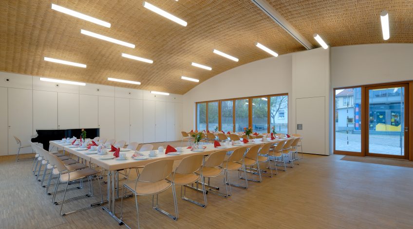 Bankettbestuhlung mit hellen Stühlen in modernem Veranstaltungsraum“ Bankettbestuhlung mit hellen Stühlen und gedeckten Tischen in einem großen Veranstaltungsraum mit Holzdecke